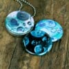 Roestvrij stalen fotomedaillon ketting met drie verschillende verwisselbare voorkanten met close up foto`s van olie druppels in water in ijsblauw en aqua blauwe tinten. Rvs/roesvrij stalen fotomedaillon. Voor het dicht bij je dragen van je favoriete foto.
