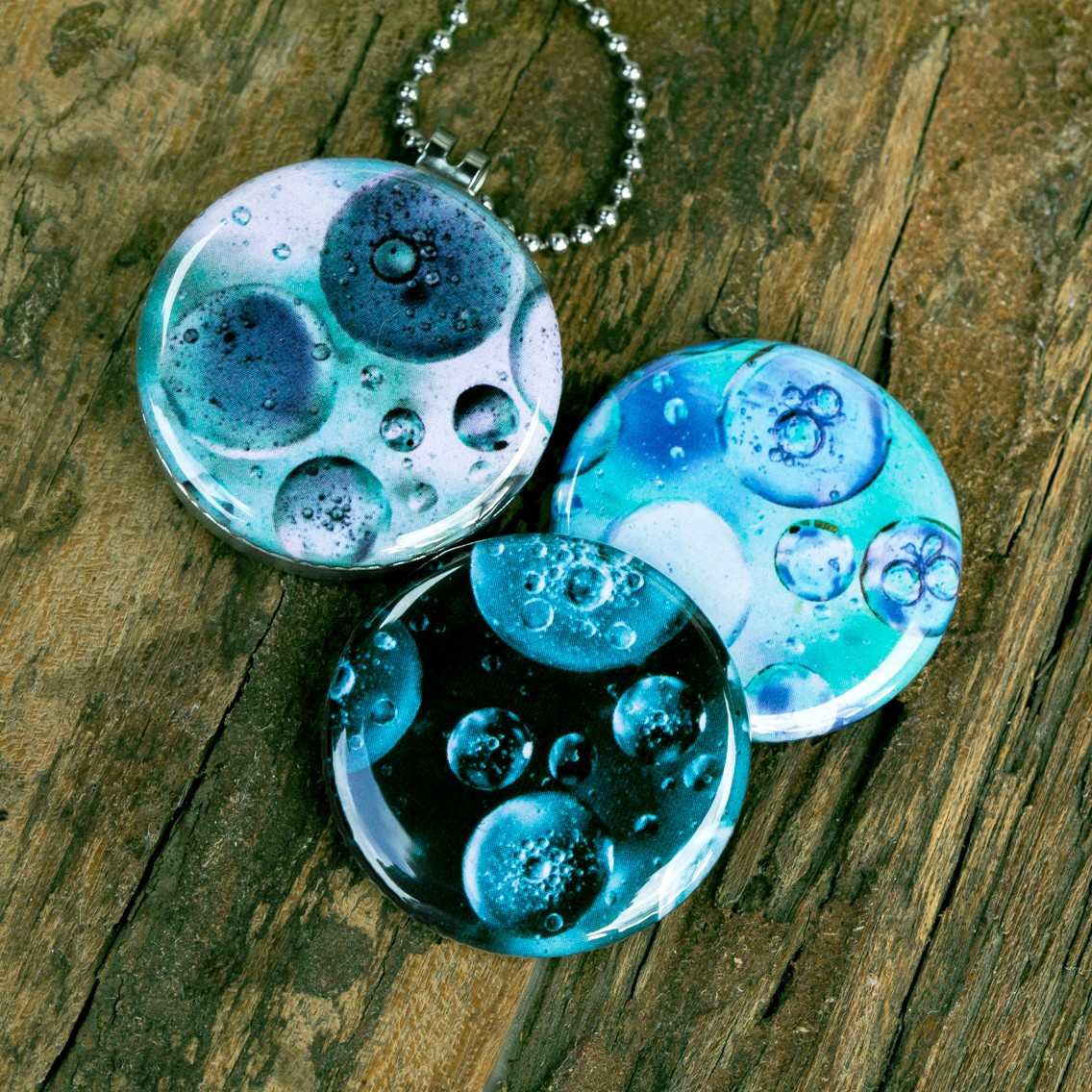 Roestvrij stalen fotomedaillon ketting met drie verschillende verwisselbare voorkanten met close up foto`s van olie druppels in water in ijsblauw en aqua blauwe tinten. Rvs/roesvrij stalen fotomedaillon. Voor het dicht bij je dragen van je favoriete foto.