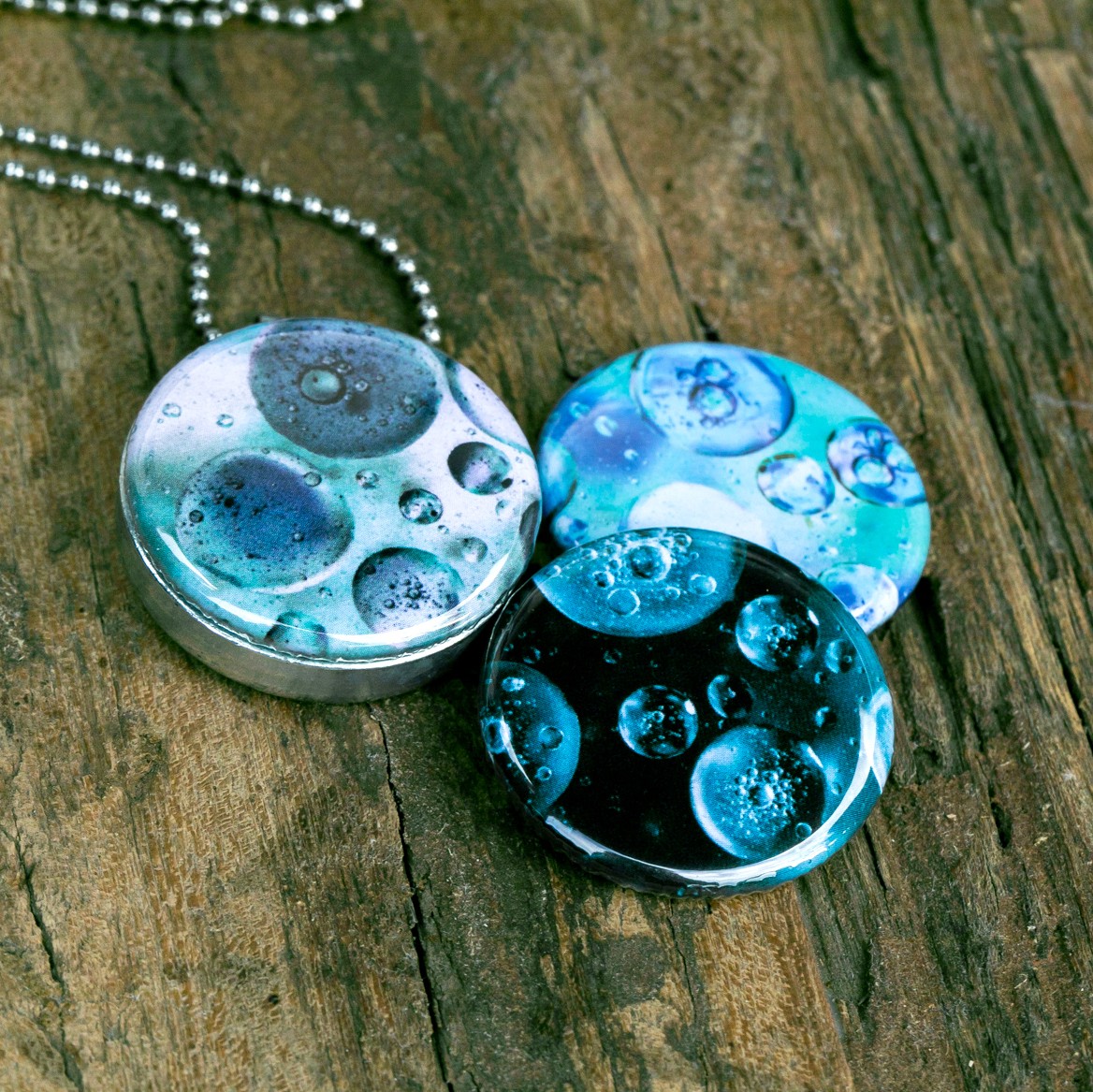 Roestvrij stalen fotomedaillon ketting met drie verschillende verwisselbare voorkanten met close up foto`s van olie druppels in water in ijsblauw en aqua blauwe tinten. Rvs/roesvrij stalen fotomedaillon. Voor het dicht bij je dragen van je favoriete foto.